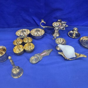 May include: A collection of brass items including a bell, a cup with a spoon, several small bowls, a multi-wick lamp, a flat dish, and a white conch shell resting on a brass stand. One brass piece features the engraved text Siddharth 2.
