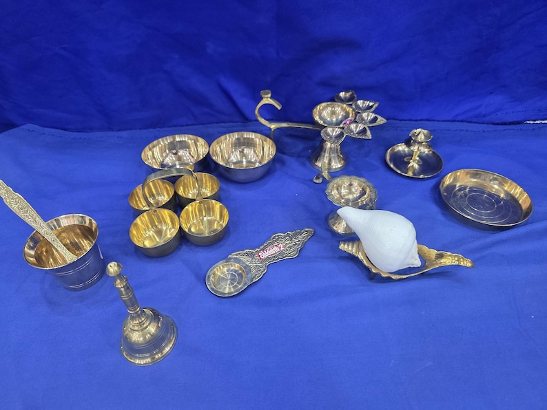 May include: A collection of brass items including a bell, a cup with a spoon, several small bowls, a multi-wick lamp, a flat dish, and a white conch shell resting on a brass stand. One brass piece features the engraved text Siddharth 2.