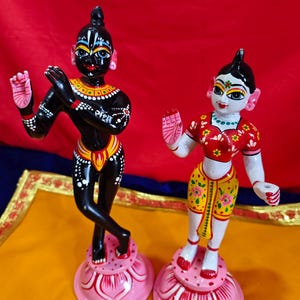 May include: Two hand-painted figurines of Hindu deities. One is black with yellow and white accents, the other is white with red and yellow clothing. Both stand on pink lotus flower bases.