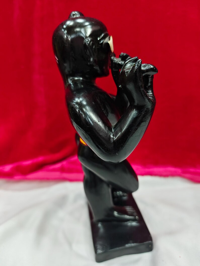 Pu&ograve; includere: Una scultura nera di una persona che suona il flauto. La figura &egrave; lucida e poggia su una base rettangolare. Lo sfondo &egrave; un tessuto rosso scuro.