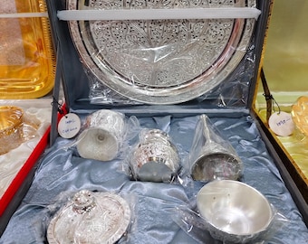 May include: A silver-colored serving set with intricate floral patterns, including a large round tray, a small bowl, a spoon, and other serving pieces, all displayed in a dark gray box with a blue satin lining.