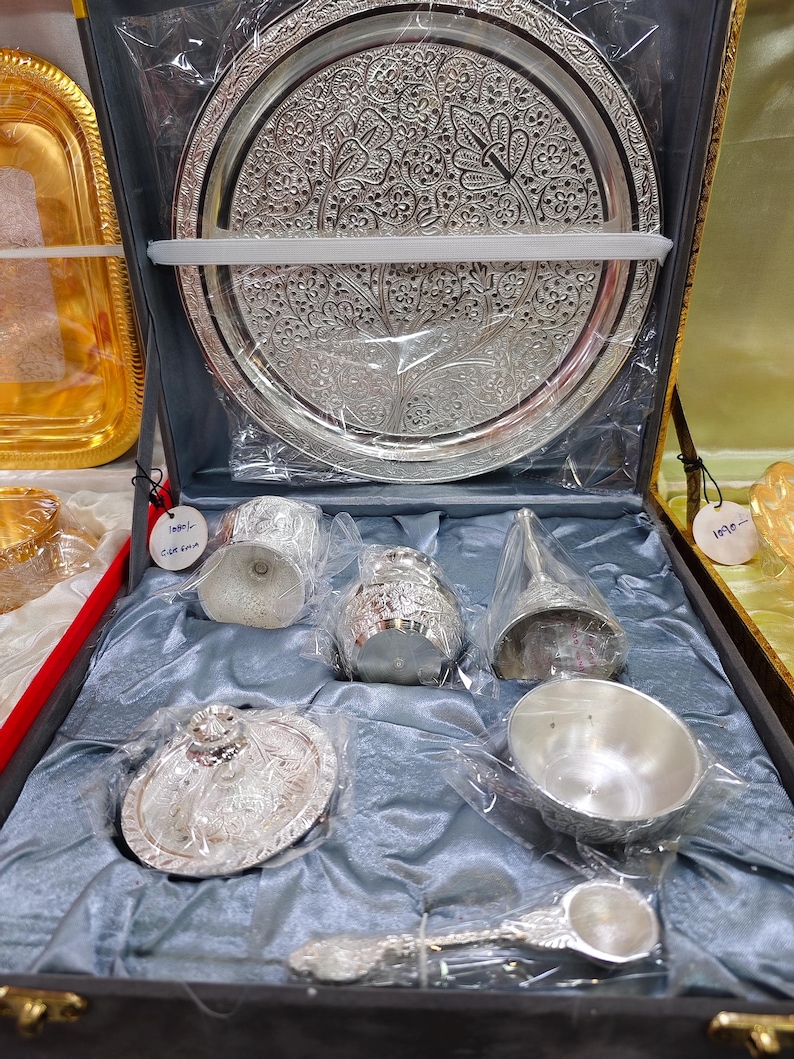 May include: A silver-colored serving set with intricate floral patterns, including a large round tray, a small bowl, a spoon, and other serving pieces, all displayed in a dark gray box with a blue satin lining.