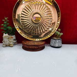 May include: A round, polished brass plate featuring a central circular indentation surrounded by radiating fluted grooves. The rim has a perforated edge design. A small rectangular sticker with the number 12 is visible in the centre of the plate.