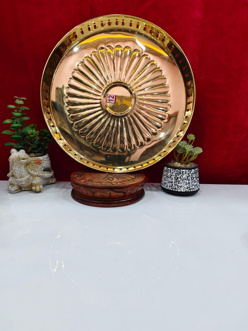 May include: A round, polished brass plate featuring a central circular indentation surrounded by radiating fluted grooves. The rim has a perforated edge design. A small rectangular sticker with the number 12 is visible in the centre of the plate.