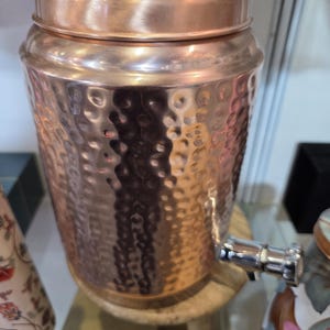 May include: A copper water dispenser with a hammered texture and a lid. The dispenser has a silver spigot and sits on a small wooden stand. The copper has a shiny, reflective surface.