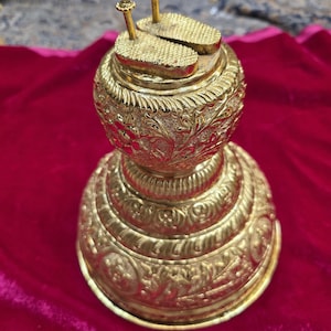 May include: A gold-colored, ornate decorative object with a round base and a detailed, textured surface. The top features two small, rectangular platforms with vertical posts. The object is likely a religious or ceremonial item.