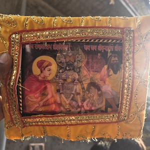 May include: A framed religious image featuring colorful illustrations of deities and figures. The image is surrounded by a yellow fabric border with gold and red embellishments. The image contains text in a foreign language.