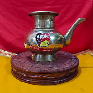 Op de afbeelding: Een messing waterpot met een tuit en een afgeronde body, op een houten standaard. De pot heeft een "Apple"-label en leest "Special Quality". De achtergrond is rood en geel.