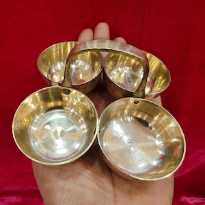Puede incluir: Un juego de cuatro cuencos pequeños y brillantes de color dorado con un asa, sostenidos en una mano sobre un fondo rojo. Los cuencos son redondos con un interior liso y un exterior ligeramente texturizado. El asa está unida a la parte superior de los cuencos.