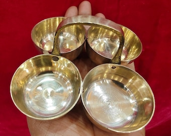 May include: A set of four small, shiny, gold-colored bowls with a handle, held in a hand against a red background. The bowls are round with a smooth interior and a slightly textured exterior. The handle is attached to the top of the bowls.