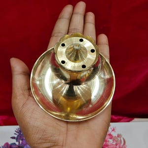 May include: A polished brass incense burner featuring a wide, circular base and a central, elevated, flower-shaped dome with five small, evenly spaced holes for holding incense sticks. The metallic surface reflects light, highlighting its smooth texture.