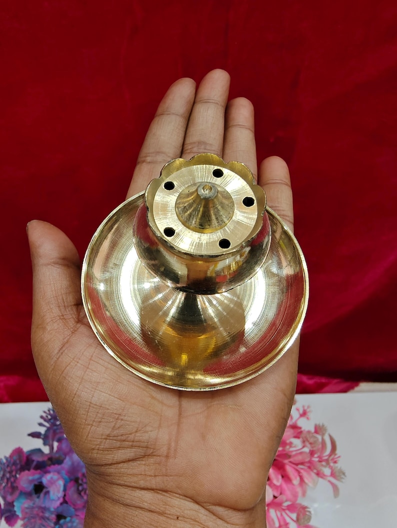 May include: A polished brass incense burner featuring a wide, circular base and a central, elevated, flower-shaped dome with five small, evenly spaced holes for holding incense sticks. The metallic surface reflects light, highlighting its smooth texture.