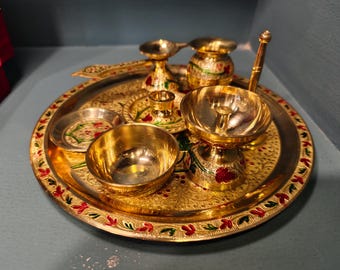 May include: A golden brass puja thali set with multiple bowls, a small pot, and a spoon. The thali and items are decorated with red, green, and gold floral patterns. The set is designed for religious ceremonies.