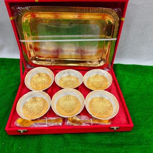May include: A red velvet gift box containing a gold-colored serving tray, six small bowls with gold-colored centers and silver rims, and two gold-colored spoons. The bowls and spoons are arranged on a red velvet base.