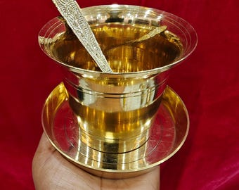 May include: A gold-coloured metal cup with a matching saucer, held in a hand. A decorative spoon rests inside the cup. The cup and saucer have a polished, reflective surface. The background is a deep red.