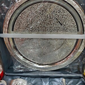 May include: Silver-colored, ornate serving tray with intricate floral and leaf patterns. The tray is round with a detailed border and is packaged in a box with other serving pieces. A white ribbon secures the tray.
