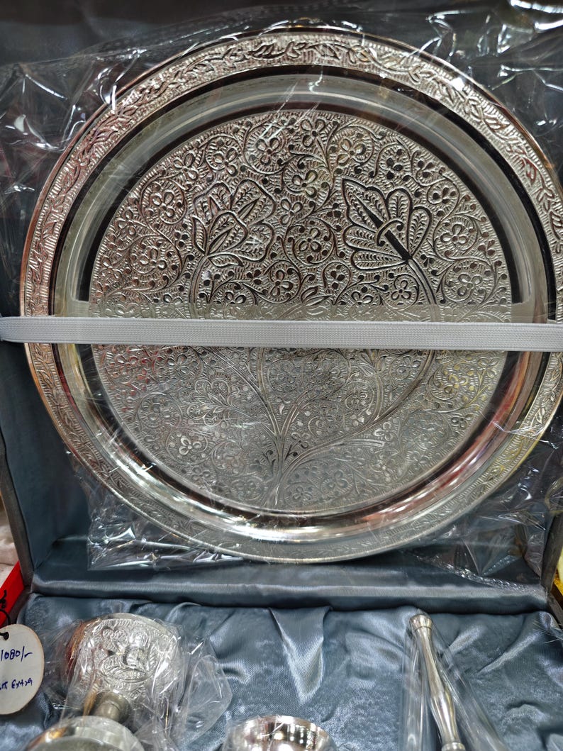 May include: Silver-colored, ornate serving tray with intricate floral and leaf patterns. The tray is round with a detailed border and is packaged in a box with other serving pieces. A white ribbon secures the tray.