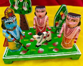 May include: A colorful, hand-painted wooden diorama featuring three figures, a cow, and two trees on a green base. The figures are dressed in traditional attire, with intricate details and vibrant colors. The cow is white with black markings.