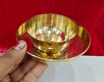 May include: A small, gold-coloured bowl and plate set. The bowl sits atop the plate, both with a shiny, metallic finish. The background is a deep red colour, and the set is resting on a white surface.