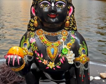 May include: A black statue of a deity with intricate gold and colorful floral detailing. The statue holds a yellow object and is adorned with jewelry. The background features a body of water and buildings.