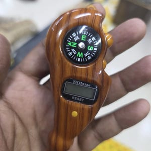 May include: A handheld wooden compass with a digital counter and the text "RESET". The compass has a black and white dial with directional markings. The digital display shows "SXH5136". The device is brown with orange accents.