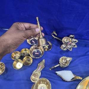 May include: A collection of brass items including a hand bell, several small bowls, a multi-wick oil lamp, a single-wick lamp, a decorative spoon-shaped vessel, and a white conch shell. Some items feature small red labels with the text DURGA.