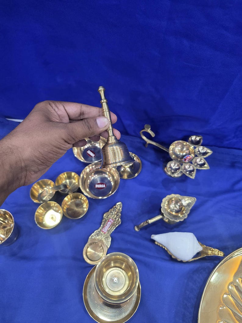 May include: A collection of brass items including a hand bell, several small bowls, a multi-wick oil lamp, a single-wick lamp, a decorative spoon-shaped vessel, and a white conch shell. Some items feature small red labels with the text DURGA.
