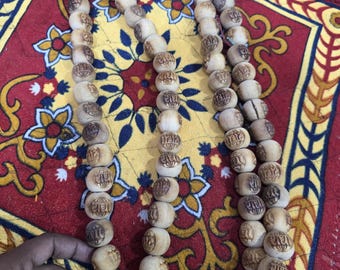 May include: Three strands of light-colored wooden prayer beads with etched symbols. The beads are resting on a red and yellow patterned textile. The beads are round and appear to be hand-carved.