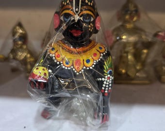May include: A black, hand-painted figurine of a deity, adorned with colorful details. The figurine is wrapped in clear plastic and features a detailed face, elaborate headwear, and decorative clothing. Other similar figurines are visible in the background.
