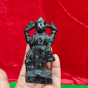 Könnte beinhalten: Kleine, schwarze Statue einer Gottheit mit goldfarbenen Augen und detaillierter, strukturierter Oberfläche. Die Figur steht mit erhobenen Armen vor rotem Hintergrund.