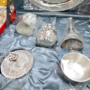 May include: A silver-colored decorative set, including a tray, bell, small bowls, and a spoon, all with intricate embossed designs. The items are packaged in clear plastic and presented on a blue satin-like fabric.