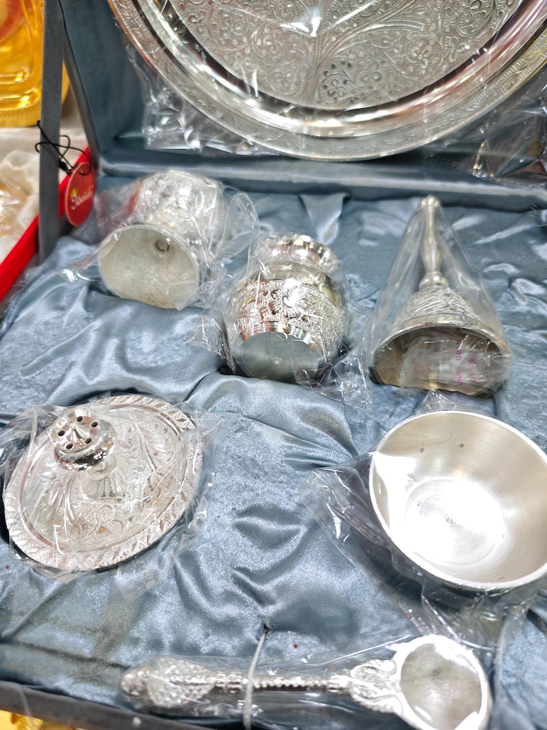 May include: A silver-colored decorative set, including a tray, bell, small bowls, and a spoon, all with intricate embossed designs. The items are packaged in clear plastic and presented on a blue satin-like fabric.