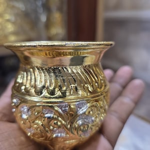 以下が含まれることがあります： 幅広の縁と丸みを帯びたボディを持つ、小さな装飾的な金色の金属製のポット。 ポットは、複雑なエンボス加工された花柄とスクロールワークのデザインが特徴で、シルバーのアクセントが付いています。 ポットは手で持たれています。