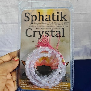 May include: A clear plastic package containing a necklace of spherical, translucent beads. The package has text that reads "Sphatik Crystal" and describes the benefits of the stone. A pink tassel is attached to the necklace.
