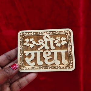 May include: A rectangular wooden plaque with the Hindi text "Shree Radha" carved in a stylized font. The plaque is framed with a decorative border and floral accents. The background is a solid red.