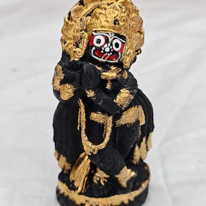 May include: A small, black figurine with gold accents. The figure has a detailed face with red and white paint and is wearing a gold headdress. The figurine is in a seated position with hands clasped together. The base is also gold.