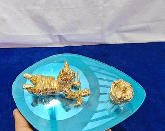 May include: A gold-colored decorative tray with a turquoise blue, teardrop-shaped design. The tray holds three gold figurines: a reclining figure, a standing figure, and a small round object. The tray is held by a hand.