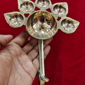 May include: A polished brass oil lamp featuring a large central circular bowl surrounded by five smaller leaf-shaped bowls arranged in a fan pattern. The lamp includes a sturdy vertical handle extending downward from the base of the central bowl.