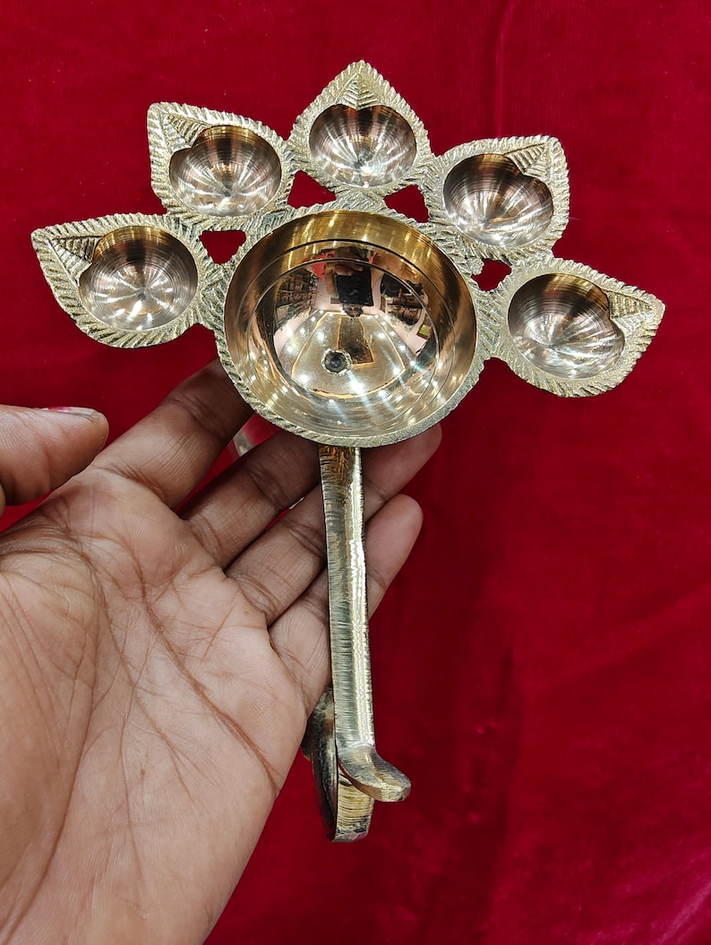 May include: A polished brass oil lamp featuring a large central circular bowl surrounded by five smaller leaf-shaped bowls arranged in a fan pattern. The lamp includes a sturdy vertical handle extending downward from the base of the central bowl.