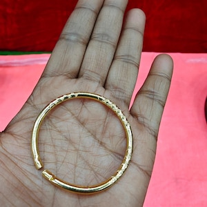 Puede incluir: Un brazalete dorado con un diseño texturizado en forma de diamante. El brazalete abierto se sostiene en una mano sobre un fondo rosa. El brazalete tiene aproximadamente 6,35 cm de diámetro.