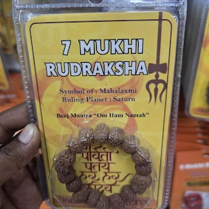 Puede incluir: Un brazalete 7 Mukhi Rudraksha empaquetado. La tarjeta amarilla insertada presenta texto que incluye "7 Mukhi Rudraksha", "Símbolo de: Mahalaxmi" y "Planeta gobernante: Saturno". El brazalete está hecho de cuentas marrones.