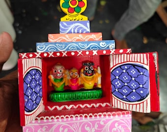May include: A colorful, hand-painted wooden shrine featuring three small deity figures inside. The shrine has a tiered design with vibrant colors, including pink, red, blue, and yellow, and decorative patterns. The figures are adorned with bright clothing and detailed facial features.