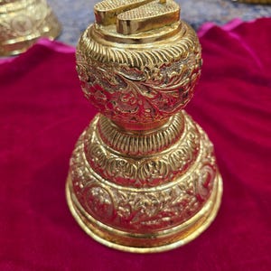May include: Ornate gold-colored decorative object with intricate floral and geometric patterns. The object has a rounded body and a tiered base, topped with two rectangular elements and two small vertical posts. The object is displayed on a red velvet surface.