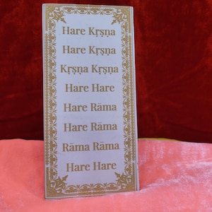 Può includere: Una carta rettangolare con testo dorato e bordi decorativi. Il testo sulla carta recita "Hare Krşņa, Hare Krşņa, Krşņa Krşņa, Hare Hare, Hare Rāma, Hare Rāma, Rāma Rāma, Hare Hare". La carta è su una superficie rosa con uno sfondo rosso.