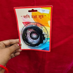 Peut inclure: Un pendentif circulaire emballé avec un cordon noir. Le pendentif est dans un étui en plastique transparent, fixé à une carte rouge et jaune avec du texte en langue étrangère. Le fond est de couleur rouge uni.