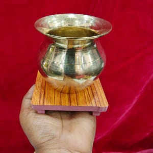 May include: A polished brass pot with a wide, flared rim and a rounded body, resting on a square, wood-grain patterned base. The metallic surface of the vessel reflects light, showcasing a smooth, reflective finish and a simple, functional design.