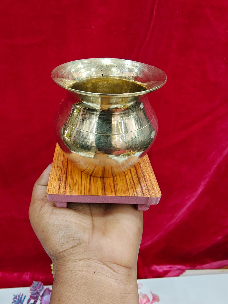 May include: A polished brass pot with a wide, flared rim and a rounded body, resting on a square, wood-grain patterned base. The metallic surface of the vessel reflects light, showcasing a smooth, reflective finish and a simple, functional design.