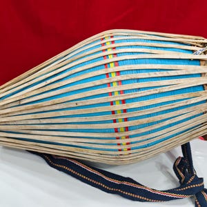 May include: A traditional Indian percussion instrument, the mridangam, with a blue body and natural wood accents. The drum is secured with woven strips and features a blue and patterned strap.