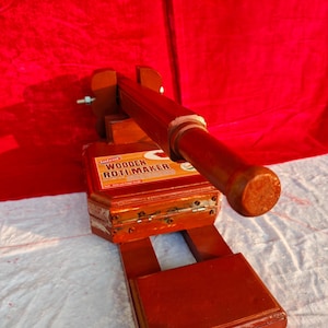 Könnte beinhalten: Ein rotbrauner Roti-Maker aus Holz, vor rotem Hintergrund. Das Gerät hat eine rechteckige Basis, einen Klappdeckel und einen langen Griff. Der Text "WOODEN ROTI MAKER" ist auf der Vorderseite zu sehen.