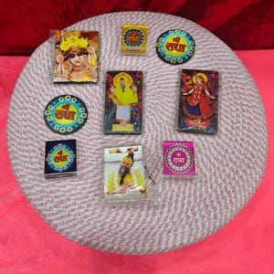 May include: A collection of colorful religious magnets and small cards featuring images of deities and the text "श्री राधा". The items are arranged on a round, woven surface with a pink and white pattern.